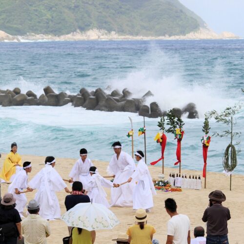 鹿児島県肝付町の岸良エリアで古くから受け継がれてきた伝統行事「夏越（なごし）どん」は、全国的には「夏越祭」と呼ばれています。平田神社に伝えられており、例年8月14日に行われます。岸良海岸での神舞奉納と茅縄くぐりが見どころです。白い砂浜と青々とした海、真っ赤な神面三体、白い衣装で神舞を奉納する舞手たちは、肝付町でしか見られない夏の風物詩です。