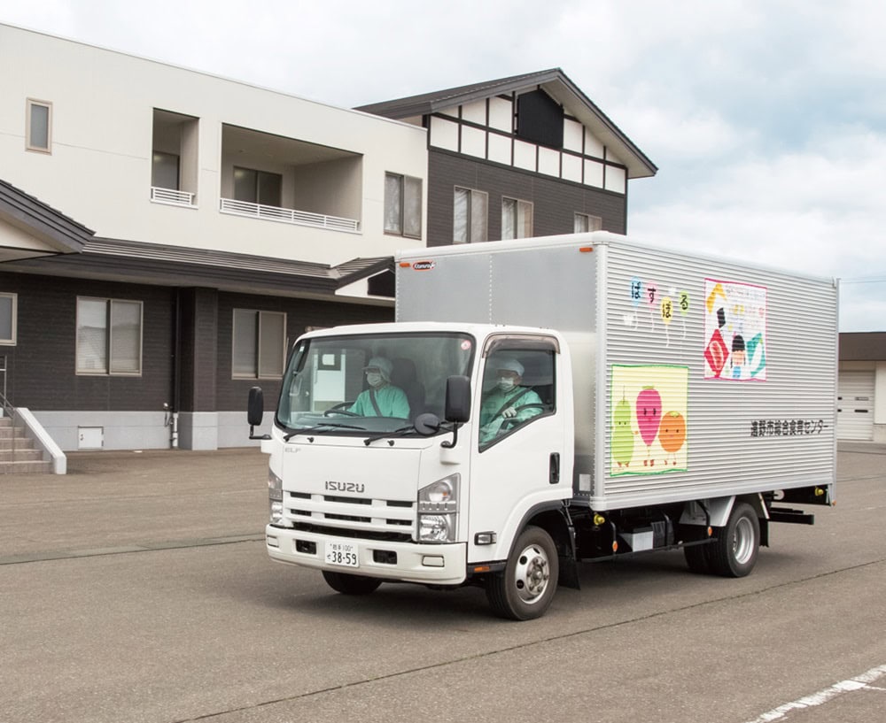 給食センターの入った“ぱすぽる”の建物の前から、できたての給食を積んだトラックが学校に向けて次々に走り出す（岩手県遠野市）