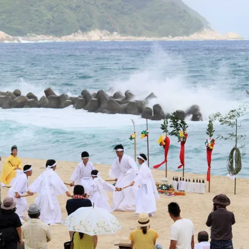 鹿児島県肝付町の岸良地区に伝わる伝統行事「岸良のナゴシドン」は、古くから人々の暮らしに根ざした夏越祭です。 平田神社を舞台に、地域住民が一体となって神楽を奉納し、一年の無病息災を祈願します。力強い太鼓の音と神楽の舞が、夏の暑さを吹き飛ばし、人々の心を清めます。白い砂浜と青々とした海、真っ赤な神面三体、白い衣装で神舞を奉納する舞手たちは、肝付町でしか見られない夏の風物詩です。