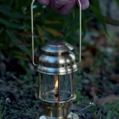 地上に置かれた真鍮製ランタン「まほろば」を男性の手が上から持ち上げようとしており、中で炎が灯っている様子。