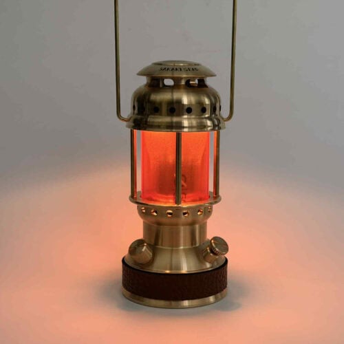 白い背景で、鮮やかなオレンジ色の光を放つ真鍮製ランタン「まほろば」が置かれ、底部に黒いレザーが巻かれている様子。